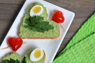 Sandwiches for breakfast. Heart-shaped bread slices smeared with ground avocado. Decorated with boiled quail eggs, parsley and tomatoes. Unfolded on a white rectangular plate.