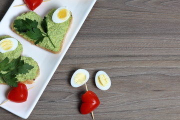 Sandwiches for breakfast. Heart-shaped bread slices smeared with ground avocado. Decorated with boiled quail eggs, parsley and tomatoes. Unfolded on a white rectangular plate.