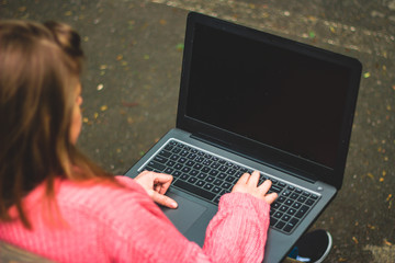 View from behind of a young blond woman in a pink blouse holding her laptop outdoors – Girl working on computer in the park – Concept image for teens using technology everywhere