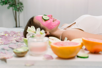 Obraz na płótnie Canvas Beautiful young woman is getting facial clay mask at spa, lying with cucumbers on eyes. Side view. Skin care, natural cosmetics concept