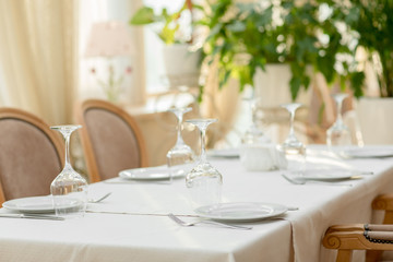 interior details of a restaurant, cafe, living room. Dining table with serving. Table with white tablecloth and glasses. Gentle beige background with green plants