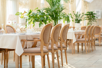 interior details of a restaurant, cafe, living room. Dining table with serving. Table with white tablecloth and glasses. Gentle beige background with green plants. restaurant chairs