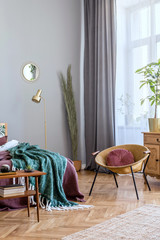 Stylish and luxury interior of bedroom with design furnitures, honey yellow armchair, gray macrame, gold lamp and elegant accessories. Beautiful bed sheets, blankets and pillows. Cozy home decor.