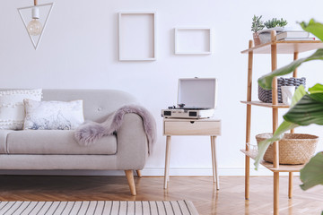 Stylish and cozy scandinavian white interior with design sofa, bookstand with accessories, pillows, blanket, gramophone and mock up photo frames. White background walls and modern triangle lamp. 