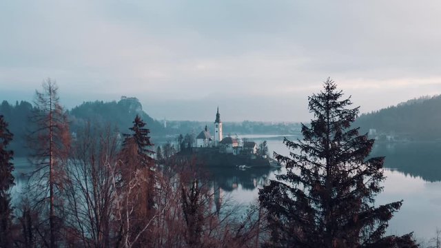 A drone shot flying over trees and approaching an island hit by first sun rays on Lake Bled in the middle of the cold winter morning. Famous tourist destination.