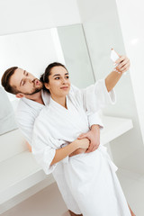 cheerful man with duck face taking selfie while hugging girlfriend in bathroom