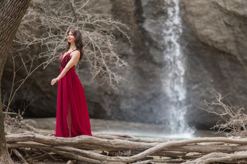 Young beautiful woman is wearing fashion red dress walking in forest near the waterfall