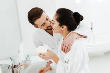 cheerful boyfriend kissing happy girl holding container with face cream