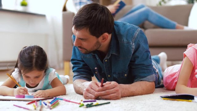 family, fatherhood and leisure concept - father spending time with his little daughters drawing in sketchbooks by crayons and lying on floor at home