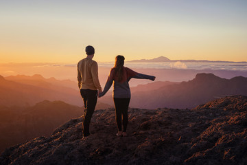 young couple from behind holding hands while standing on mountain top with scenic landscape and...