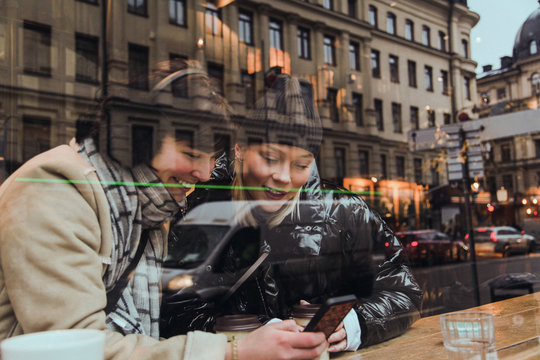 Smiling female friends using mobile phone while sitting in cafe seen through window