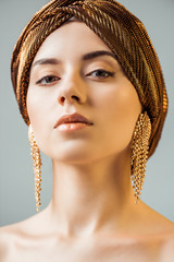 young naked woman with shiny makeup, golden rings in turban looking at camera isolated on grey