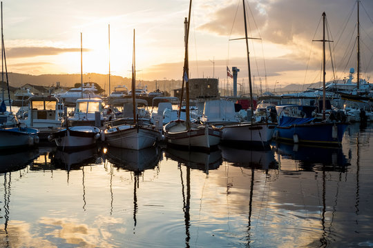 Sailboats moored in harbour reflected in calm sea at sunset.