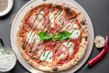 Pizza Margherita with green salsa and leaf of basyl on wooden plate, garlic, red pepper and bowls with flour and spices near. Black background, top view, flat lay, vertical image. Natural light.