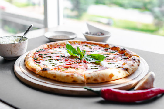 Pizza Margherita on wooden plate with red pepper and white bowl near, everything on the table near the window. Natural light. Copy space. Horizontal image.