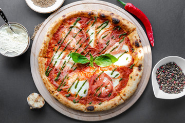 Pizza Margherita with green salsa and leaf of basyl on wooden plate, garlic, red pepper and bowls with flour and spices near. Black background, top view, flat lay, horizontal image. Natural light. 
