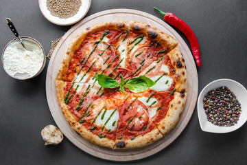 Pizza Margherita with green salsa and leaf of basyl on wooden plate, garlic, red pepper and bowls with flour and spices near. Black background, top view, flat lay, horizontal image. Natural light. 