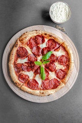 Pepperoni pizza with leaf of basyl on the wooden plate, cumin and bowl with flour near. Top view, flat lay, black background. Copy space, vertical image. Natural light. 
