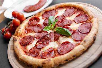Close up Pepperoni pizza with basyl leaf in the middle on the wooden plate, plate with salami slices, cherry tomato branch near. Black background. Horizontal image. Natural light. 