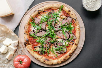 Pizza with roast beef, arugula on the wooden plate. Tomato and pieses of cheese near on the table. Horizontal. Natural light. Black background. Flat lay, top view.