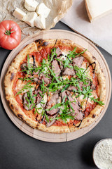 Pizza with roast beef, arugula on the wooden plate. Tomato and pieses of cheese near on the table. Vertical. Natural light. Black background. Flat lay, top view.