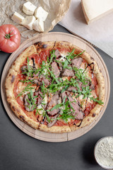 Pizza with roast beef, arugula on the wooden plate. Tomato and pieses of cheese near on the table. Vertical. Natural light. Black background. Flat lay, top view.