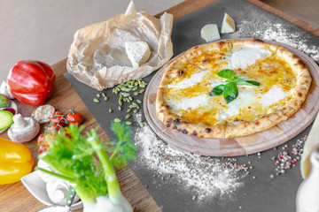 Pizza with four kinds of cheese on the table in the restaurant. Spices, chesse and different vegetables near. Horizonal image. Natural light. Black background. 