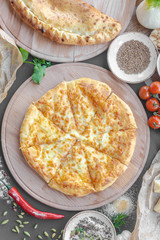 Georgian khachapuri pie with cheese on wooden plate, cheese, red pepper, tomatoes, mushrooms, herbs, onion and bowls with spices nearby on the table. Horizontal image. Top view, flat lay.
