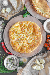 Georgian khachapuri pie with cheese on wooden plate, cheese, red pepper, tomatoes, mushrooms, herbs, onion and bowls with spices nearby on the table. Horizontal image. Top view, flat lay.
