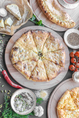 Georgian khachapuri pies with cheese and meat on the plate, red pepper, tomatoes, mushrooms, herbs, onion and bowls with spices nearby on the table. Vertical image. Top view, flat lay.