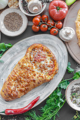 Georgian khachapuri pie with cheese and meat on the plate, red pepper, tomatoes, mushrooms, herbs, onion and bowls with spices nearby. Black background. Vertical image.  Top view, flat lay.