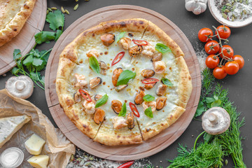 Pizza with sea food, shrimps, mussels, cheese on wooden deck, red pepper, tomatoes, mushrooms, herbs, onion, bowl with spices nearby on the table. Horizontal image. Top view, flat lay. Natural light. 