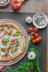 Pizza with sea food, shrimps, mussels, cheese on wooden deck, tomatoes, mushrooms, herbs, bowl with spices nearby on the table. Vertical image. Top view, flat lay. Natural light. 