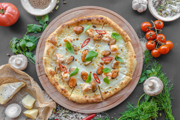 Pizza with sea food, shrimps, mussels, cheese on wooden deck, red pepper, tomatoes, mushrooms, herbs, onion, bowl with spices nearby on the table. Horizontal image. Top view, flat lay. Natural light. 