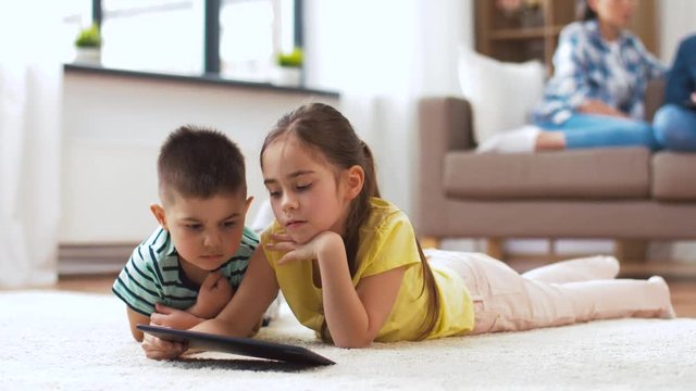 childhood, technology and family concept - little brother and sister with tablet pc computer lying on floor at home