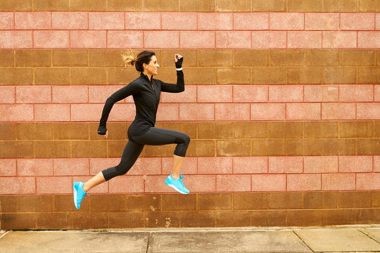 A female runner jumping next to a red cinderblock wall.