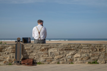 Back view of man with laptop and longboard sitting on a wall in front of beach and sea looking at distance