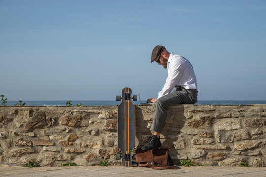 Man sitting on a wall in front of the sea working on laptop