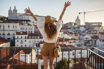 Blonde woman standing on the balcony and looking at coast view of the southern european city with sea during the sunset, wearing hat, cork bag, safari shorts and white shirt - obrazy, fototapety, plakaty