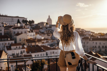 Blonde woman standing on the balcony and looking at coast view of the southern european city with sea during the sunset, wearing hat, cork bag, safari shorts and white shirt