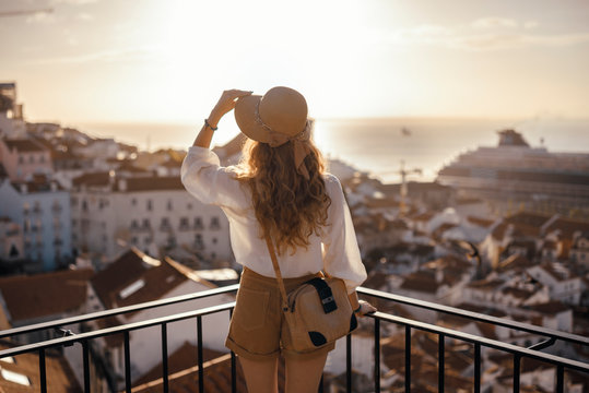 Blonde woman standing on the balcony and looking at coast view of the southern european city with sea during the sunset, wearing hat, cork bag, safari shorts and white shirt