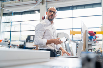 Businessman with laptop holding workpiece in factory