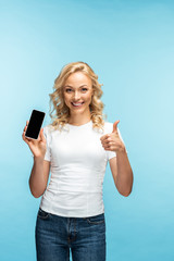 Obraz premium cheerful blonde woman showing thumb up while holding smartphone with blank screen on blue