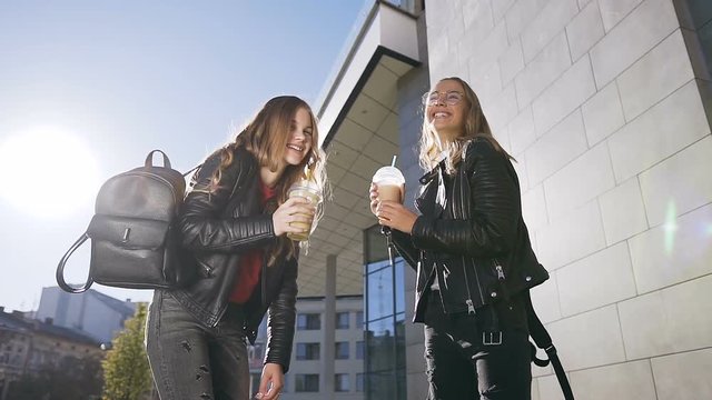 Two happy female best friends walking in warm sunny day and drinking refreshing cocktail with ice on a background building in the urban city. Outdoors, lifestyle