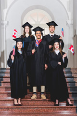 cheerful students in graduation gowns holding flags of different countries