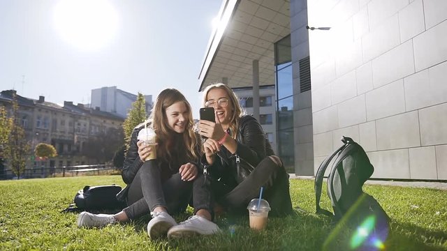 Attractive teen girlfriends with smartphone laughing at fun pictures through the internet sitting outdoors in the park in sunny weather. Outdoors