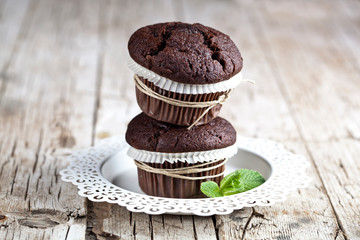 Two fresh dark chocolate muffins with mint leaves on white plate on rustic wooden table