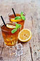 Cold iced tea with lemon, mint leaves and ice cubes in two glasses on rustic wooden table background.
