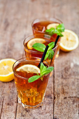 Cold iced tea with lemon, mint leaves and ice cubes in three glasses on rustic wooden table.