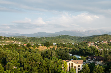 Almaty city in summer. Among the green trees are visible tall buildings and urban buildings. On the background are visible high peaks of the mountains. View from bird flight.